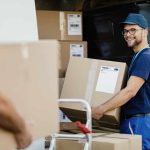 young-happy-manual-worker-carrying-cardboard-boxes-delivery-van-while-communicating-with-his-colleagues-scaled